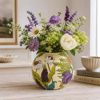 A ceramic vase with a cream background and a botanical illustration pattern, filled with a colourful flower arrangement, set on a light wooden table in a bright room.