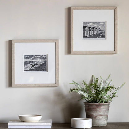 Two framed black and white landscape art prints with light wood frames and white mats, displayed on a beige wall above a shelf with books, a bowl, a candle, and a potted plant.