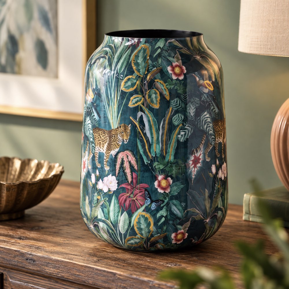 A tall ceramic vase with a dark teal and green tropical jungle print featuring leaves, flowers, and leopards, displayed on a wooden table in a styled room setting.