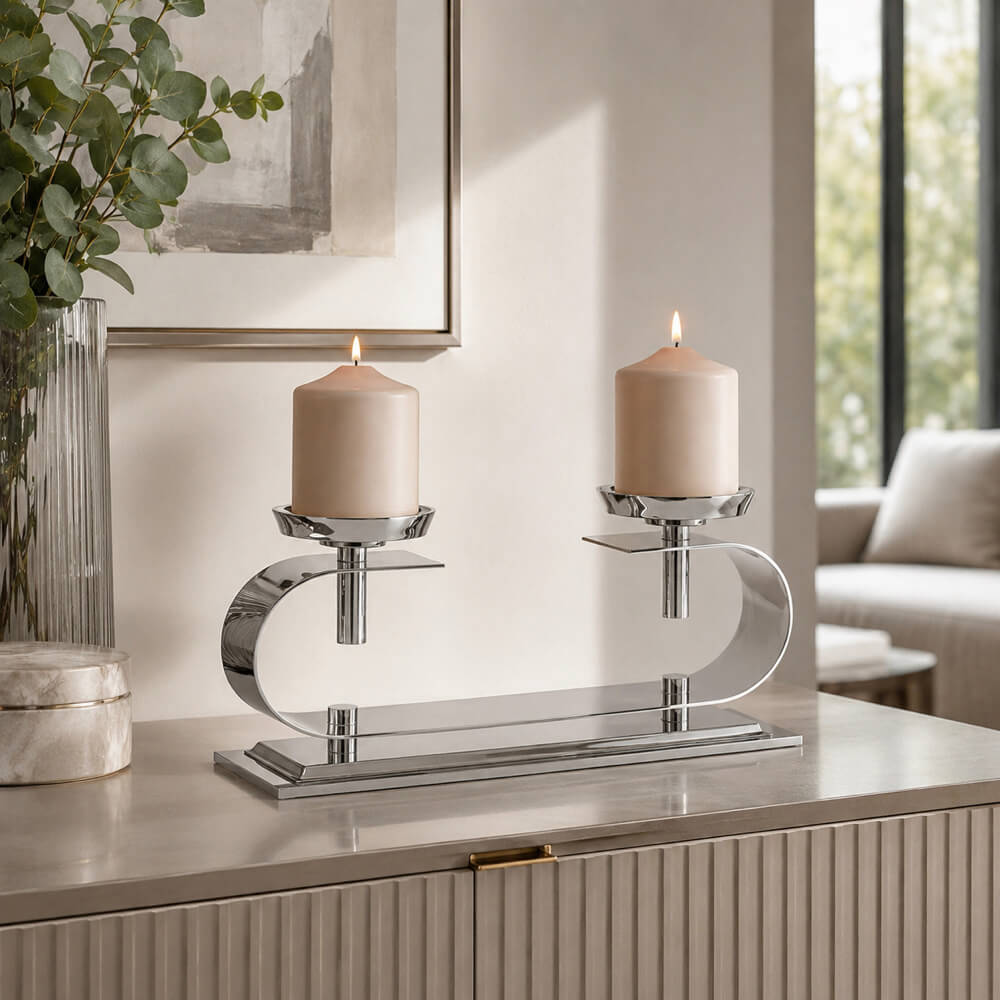 A modern two-light candle holder with a polished silver chrome base and curved arms, holding two lit beige pillar candles, set on a light gray console table in a styled living room.