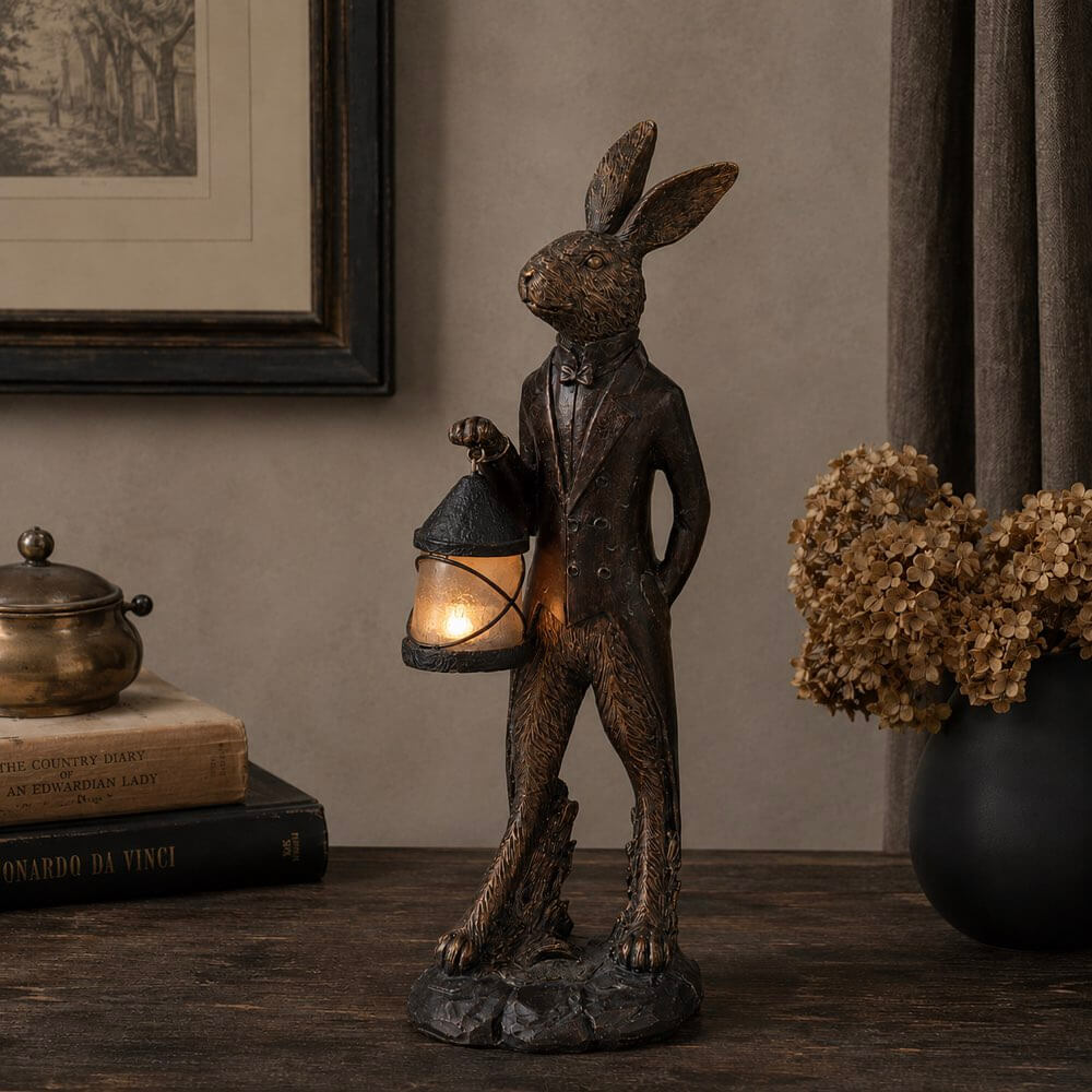 A dark bronze-finished rabbit figurine dressed in a tuxedo and holding a lit lantern, displayed on a wooden table in a styled interior setting with books, a brass object, and dried flowers.