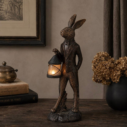 A dark bronze-finished rabbit figurine dressed in a tuxedo and holding a lit lantern, displayed on a wooden table in a styled interior setting with books, a brass object, and dried flowers.