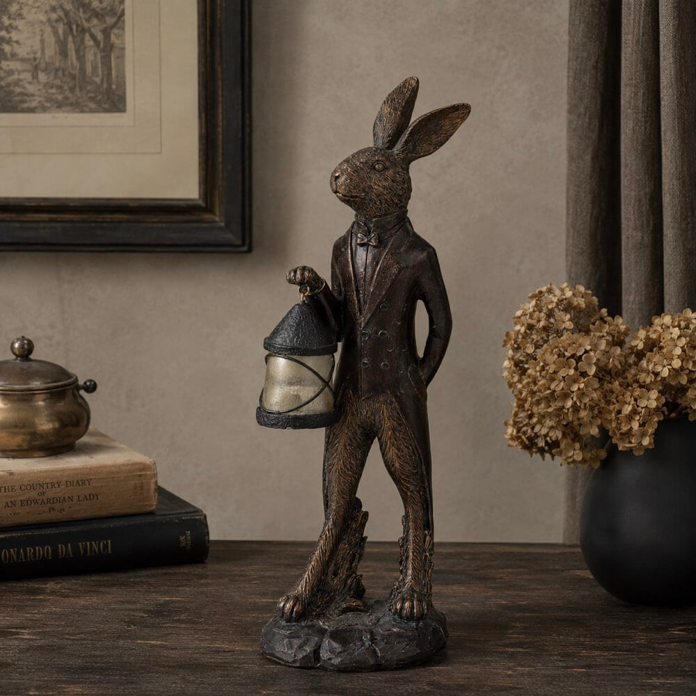 A dark bronze-finished rabbit figurine wearing a tuxedo and holding a lantern, displayed on a wooden table in a styled interior setting with books, a vase of dried flowers, and framed wall art.