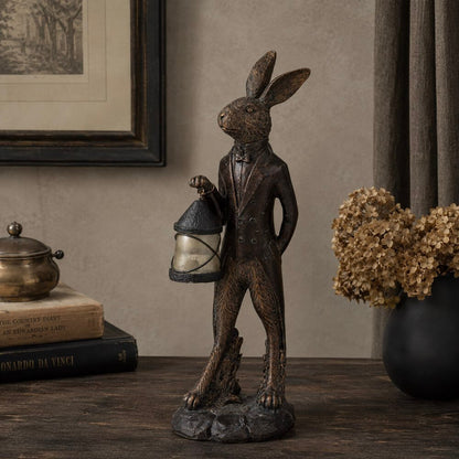 A dark bronze-finished rabbit figurine wearing a tuxedo and holding a lantern, displayed on a wooden table in a styled interior setting with books, a vase of dried flowers, and framed wall art.