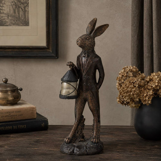 A dark bronze-finished rabbit figurine wearing a tuxedo and holding a lantern, displayed on a wooden table in a styled interior setting with books, a vase of dried flowers, and framed wall art.