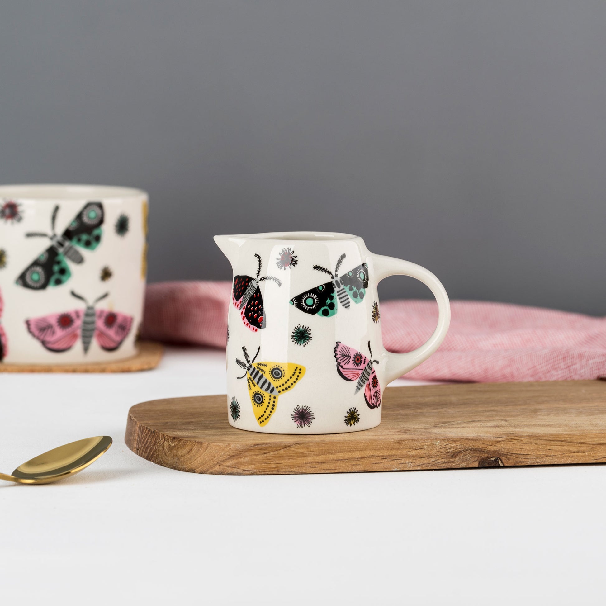 A ceramic jug with a moth pattern, placed on a wooden board next to a pink cloth, with a cup in the background that also features a moth design.
